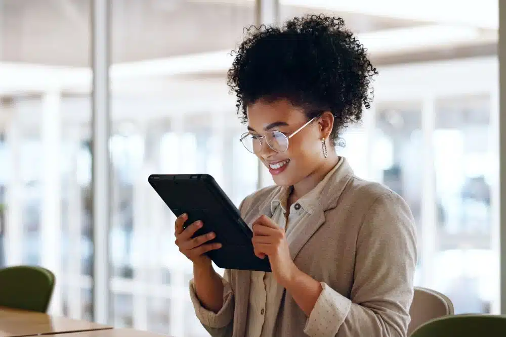 Smiling woman wearing glasses uses a tablet in a bright office setting.