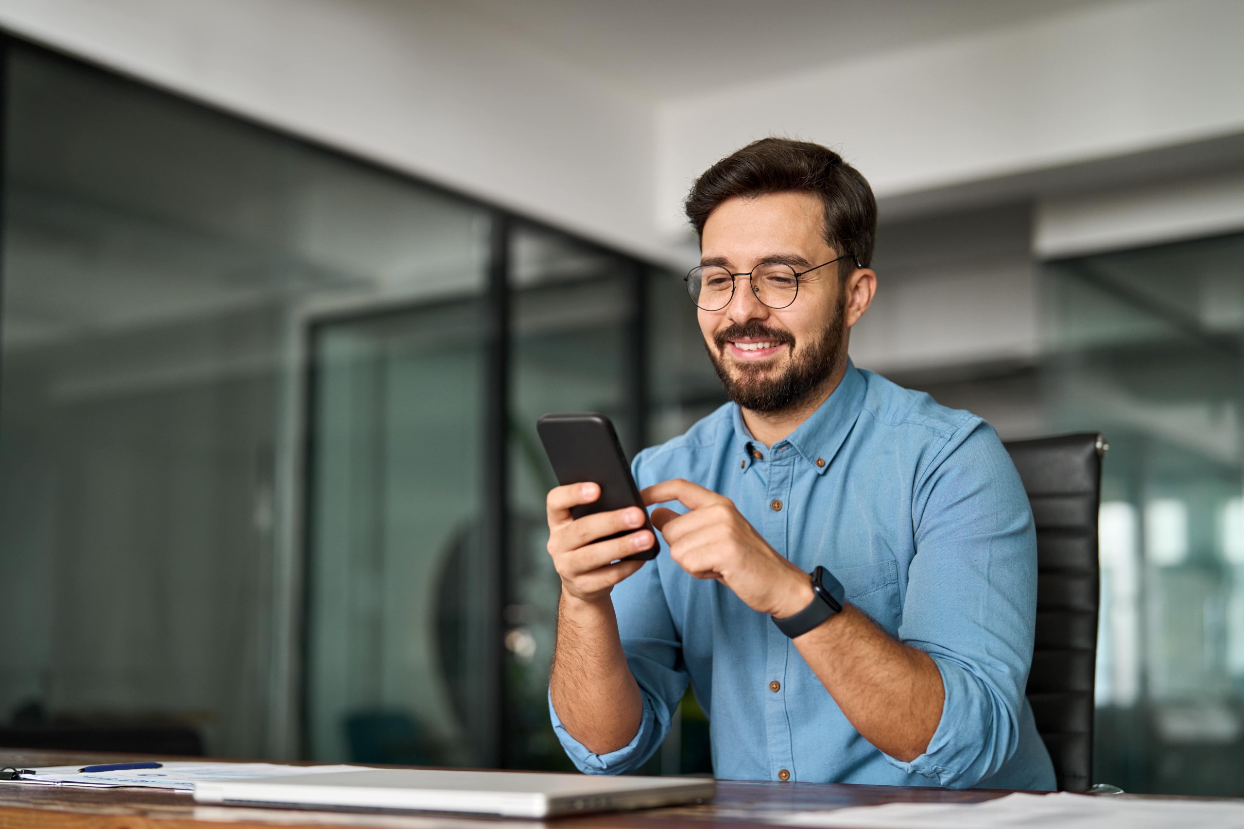 Smiling man in an office using a smartphone, suggesting communication or productivity on the go.