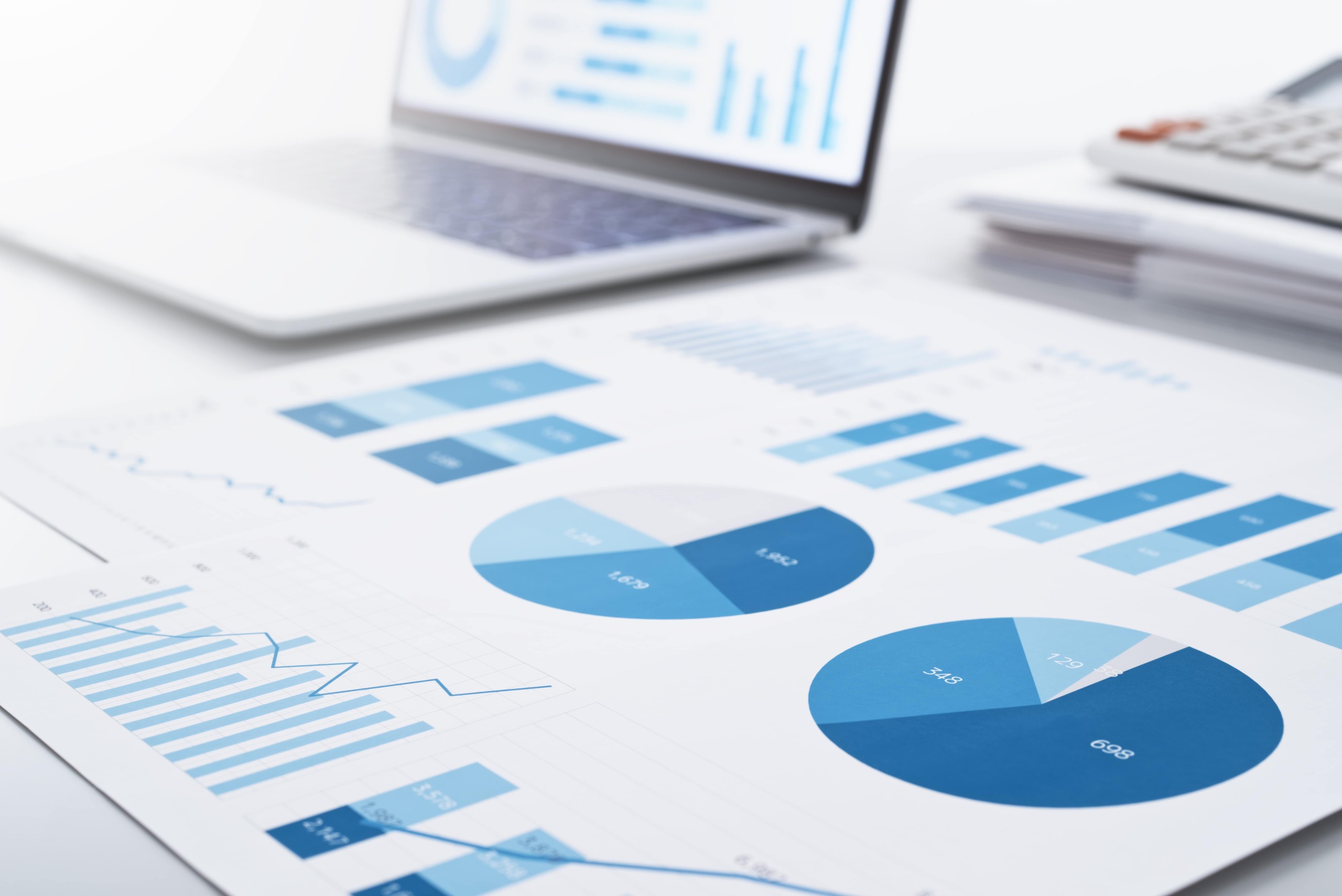 Printed financial charts and graphs on a desk with a laptop in the background, representing data analysis or reporting.