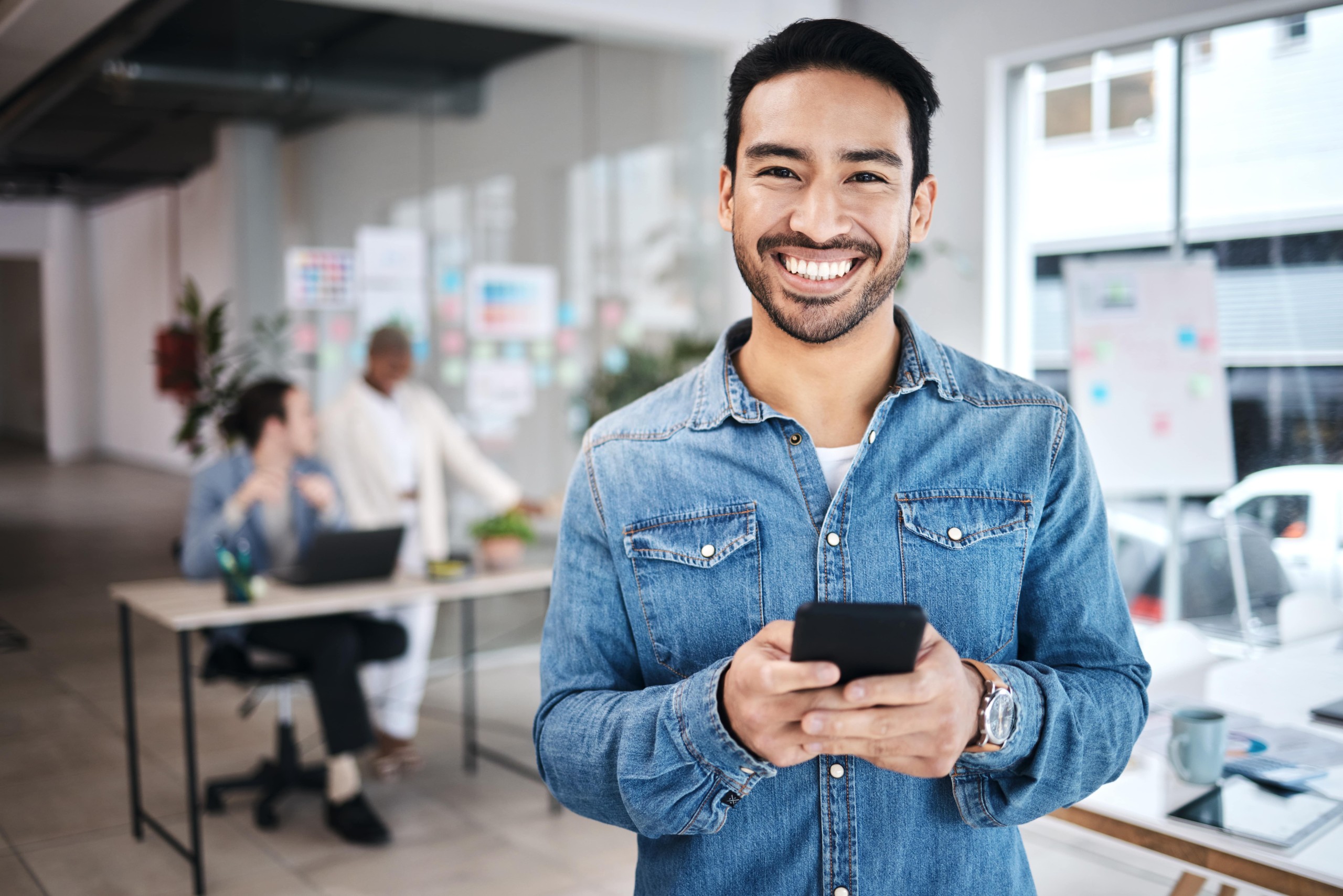 Smiling man holding a smartphone in a modern office, with colleagues collaborating in the background.