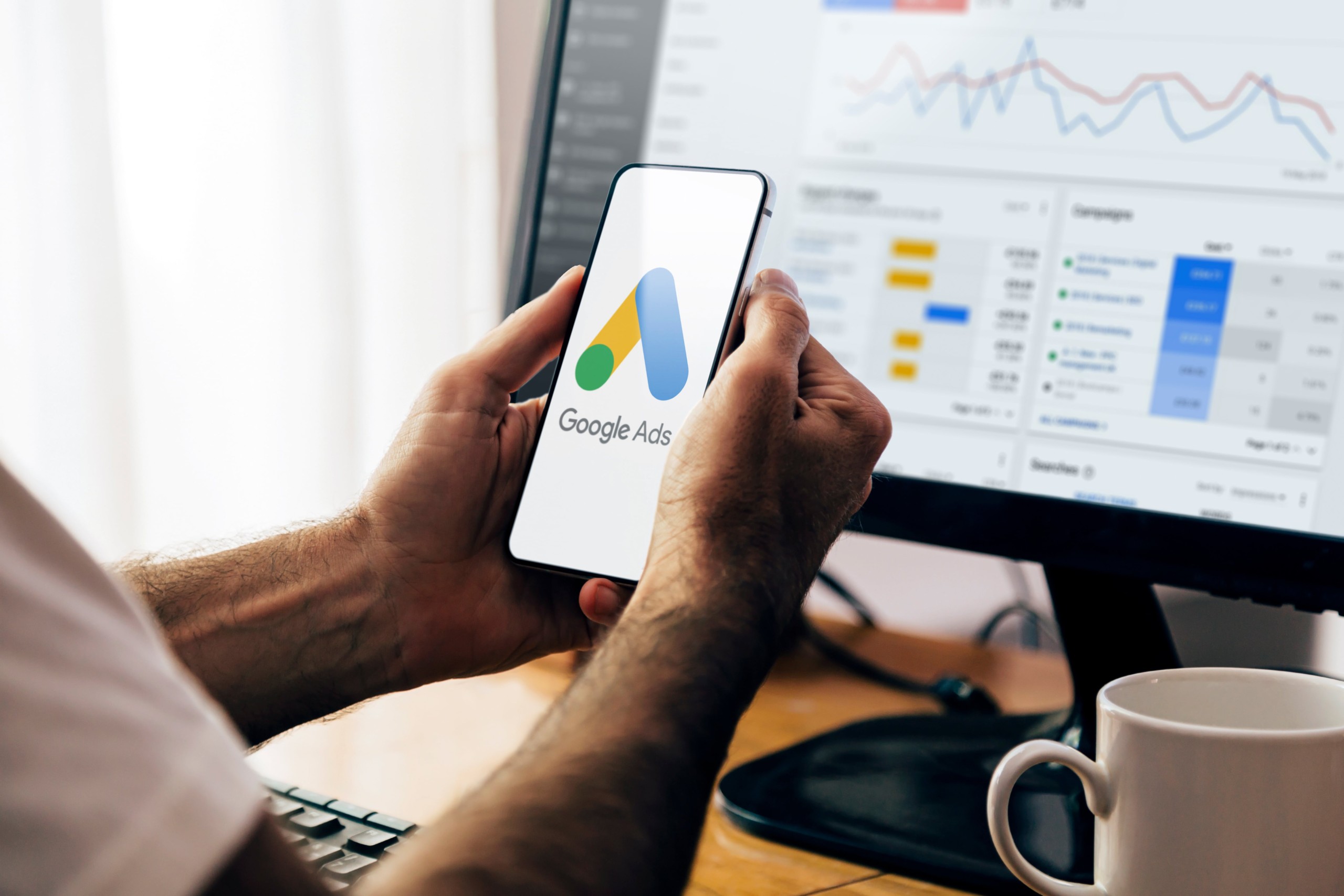 AdsServices-min Person holding a phone displaying the Google Ads logo in front of a monitor showing digital ad campaign analytics.