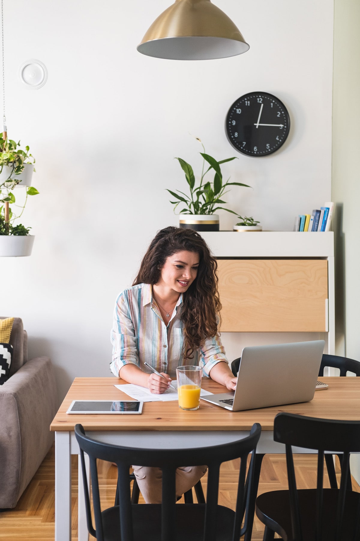 Woman working at a laptop in a bright home office, with a notepad, tablet, and glass of orange juice on the table.